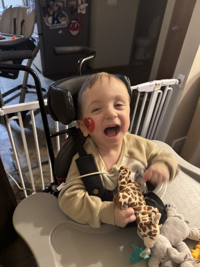 A smiling child in a wheelchair, holding a plush giraffe toy. The child has a medical tube on their face and is in a warmly lit room, exuding joy and happiness.