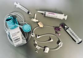 Medical supplies on a gray surface include syringes, g-tube tubing, bag for tube feeding, and connectors. Items are neatly arranged, suggesting clinical preparation.