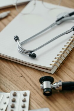 Stethoscope on an open spiral notebook, with blister packs of pills nearby, resting on a wooden table. The scene suggests a medical or clinical setting.